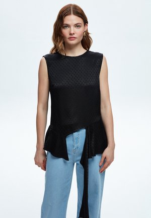 Schwarzes ärmelloses Top mit strukturierten Pünktchen, einem Peplum-Saum und einem seitlichen Bindeband, kombiniert mit hellblauen weiten Jeans.