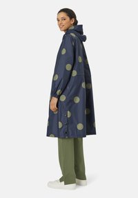 Marineblå regnfrakke med store olivengrønne prikker, der har en hætte, elastiske manchetter og trykknapper i kanten. Sammen med grønne bukser.