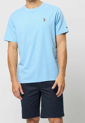Mężczyzna ubrany w jasnoniebieski t-shirt z krótkim rękawem i małym logo na piersi oraz ciemnoniebieskie szorty do kolan stoi na jednolitym tle.