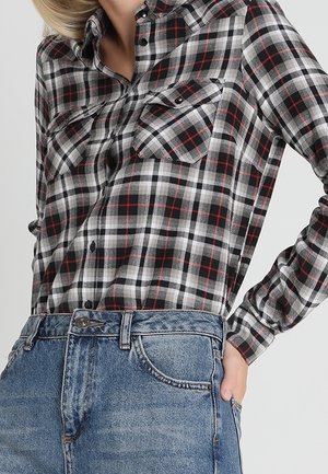 Kobieta ubrana w czarno-biało-czerwoną koszulę w kratę, wpuszczoną do wysokich, niebieskich dżinsów, z ręką opartą na biodrze.