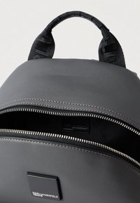 Γκρι σακίδιο φτιαγμένο από υφασμάτινο υλικό, με λαβή στο πάνω μέρος, κλείσιμο με φερμουάρ και λογότυπο μπροστά.