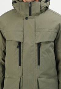 Olivgrüne Kapuzenjacke mit Reißverschluss, zwei schwarzen Reißverschlusstaschen auf der Brust und verstellbaren Kordeln. Mit Branding auf der Brust.