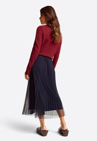 Red fuzzy sweater paired with a pleated navy midi skirt, complemented by brown block-heeled mules; shown from the back.