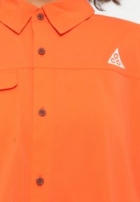 Camicetta arancione a maniche corte, realizzata in tessuto liscio, con chiusura frontale a bottoni, una tasca applicata e un piccolo triangolo bianco con logo sul petto.