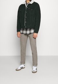 Mies, joka wear musta vihreä sherpa-vuorattu takki, musta ja valkoinen ruudullinen paita, vaaleat housut ja valkoiset lenkkitossut mustilla raidalla.