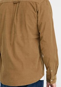 Brown corduroy shirt with long sleeves, featuring a back pleat, buttoned cuffs, and a curved hem. Soft texture and ribbed pattern visible.