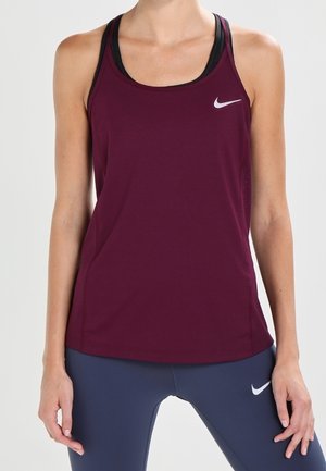 Femme portant un haut de sport sans manches bordeaux Nike et des leggings bleu marine avec logos Nike, debout, les bras relâchés le long du corps.