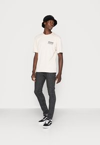 Camiseta bege clara com um pequeno gráfico preto, combinada com jeans skinny escuros e um chapéu preto tipo bucket. Ténis em preto e branco completam o visual.