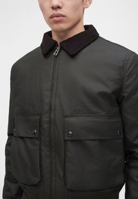 Homme portant une veste zippée vert foncé avec un col en velours côtelé noir et deux grandes poches à rabat à l'avant avec des boutons métalliques.