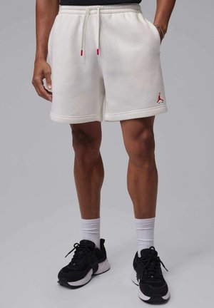 Mand iført hvide Jordan-shorts med snøre, sorte sneakers og hvide sokker, står mod grå baggrund.