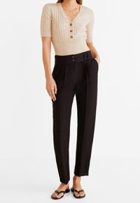 Haut beige à côtes, à manches courtes, avec un col en V et trois boutons marron, associé à un pantalon noir taille haute et des tongs noires.