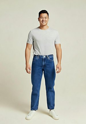 Homme debout, souriant, portant un T-shirt gris clair, un jean bleu et des baskets blanches sur un fond beige uni.