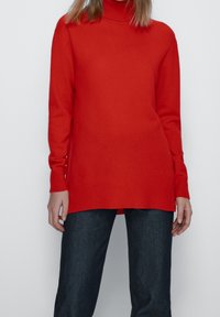 Pull à col roulé rouge en tissu tricoté doux et lisse. Manches longues et coupe décontractée avec un ourlet côtelé. Associé à un jean foncé.