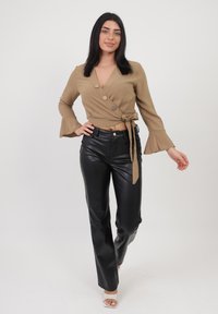 Top avvolgente beige con tre bottoni marroni e maniche a campana, abbinato a pantaloni in pelle nera a vita alta e décolleté trasparenti con cinturini.