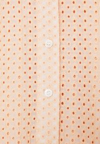 Nahaufnahme eines Stoffs mit einem Pfirsich-Hintergrund, bestickt mit kleinen Gänseblümchen-Muster mit orangefarbenem Zentrum und zwei weißen Knöpfen in der Mitte.