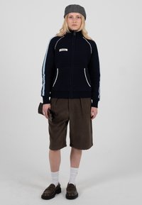 Veste en maille bleu marine avec accents blancs et fermeture éclair, associée à un short marron et des chaussettes. Le mannequin porte une casquette grise et tient des gants marron.