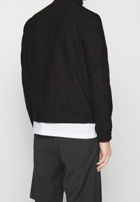 Sort bomberjakke med en glat overflade, udstyret med elastiske manchetter og en taljelængde-design, båret over en hvid T-shirt.