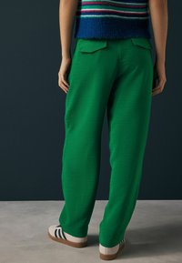 Groene broek van een gladde stof met een ontspannen pasvorm, voorzien van achterzakken en gecombineerd met een gestreepte blauwe en roze gebreide top en sneakers.