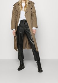 Długi beżowy trencz z epauletami, noszony na białym swetrze, zestawiony z szerokimi czarnymi skórzanymi spodniami oraz czarnymi sznurowanymi botkami.