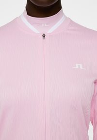 Ljusrosa sportjacka med strukturerat vågigt mönster, hög krage och vita detaljer. Har en framknäppning med dragkedja och en liten logotyp på bröstet.