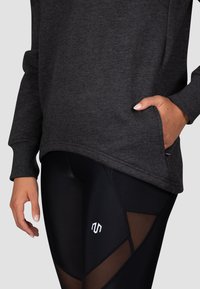 Dunkelgrauer Sweatshirt mit einem gerippten Kragen und Seitentasche; kombiniert mit schwarzen Leggings mit Mesh-Einsätzen und einem weißen Logo.
