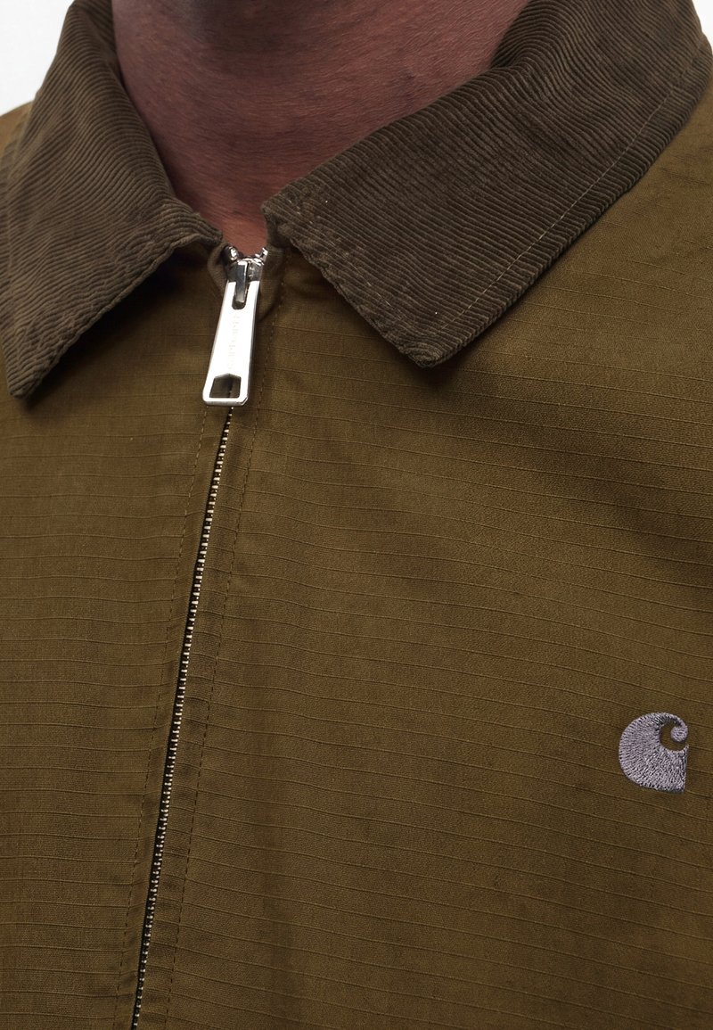 Gros plan d'une fermeture éclair fermée sur une veste marron avec un col en velours côtelé et un petit logo brodé sur la poitrine.