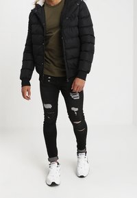 Černá péřová bunda s kapucí, olivově zelené tričko, potrhané černé skinny džíny a bílé tenisky. Texturovaný, prošívaný design s umělou kožešinou.