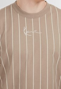 Brown striped T-shirt featuring thin white vertical stripes and a stitched cursive logo in white on the chest. Standard crew neck design.