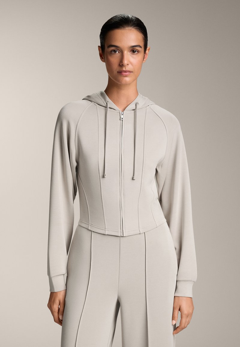 Felpa grigia chiara corta con zip anteriore, cappuccio con coulisse e maniche lunghe con polsini a coste. Materiale liscio e elasticizzato.