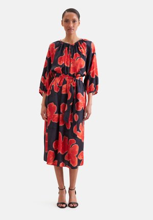 Vrouw staand, draag een zwarte midi-jurk met grote rode bloemenpatronen, driekwart mouwen en een gestrikte tailleband, gecombineerd met zwarte hakken.