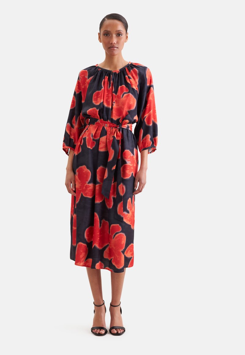 Femme debout portant une robe midi noire avec de grands motifs floraux rouges, manches trois-quarts et ceinture nouée à la taille, associée à des talons noirs.