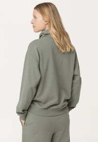 Felpa verde oliva con colletto alto e ripiegato, vestibilità comoda, polsini e orlo a coste. Tessuto morbido. Indossata con pantaloni abbinati.