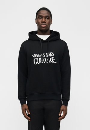 Mladi moški, oblečen v črno kapuco Versace Jeans Couture in črne hlače, stoji pred enostavnim svetlim ozadjem.