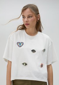 Camiseta blanca de algodón con un corte crop, que presenta múltiples adornos en forma de ojo de varios tamaños y diseños coloridos.