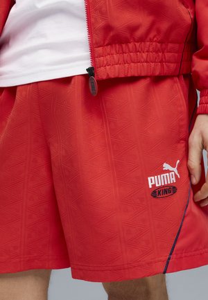 Czerwone szorty Puma King z subtelnym geometrycznym wzorem, noszone z dopasowaną czerwoną kurtką i widoczną białą koszulką pod spodem.