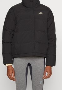 Svart puffjacka med hög krage, quiltad textur och broderad logo på vänster bröst. Matchad med grå träningsleggings.