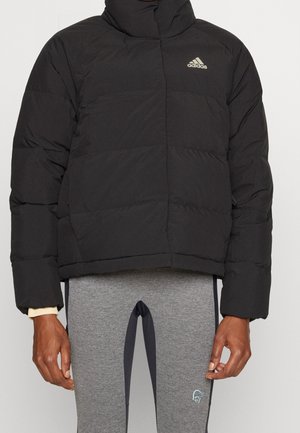 Doudoune noire avec un col haut, texture matelassée et logo brodé sur la poitrine gauche. Associée à des leggings de sport gris.