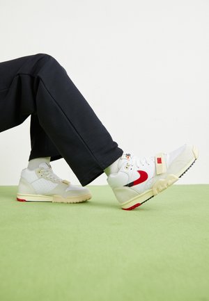 White and cream sneakers with a red accent, featuring mesh and suede materials, textured detailing, and a strap across the front.
