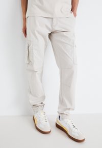 Osoba nosząca jasnobeżowe spodnie cargo i białe sneakersy z żółtymi akcentami, stojąca na tle jednolitej białej ściany.