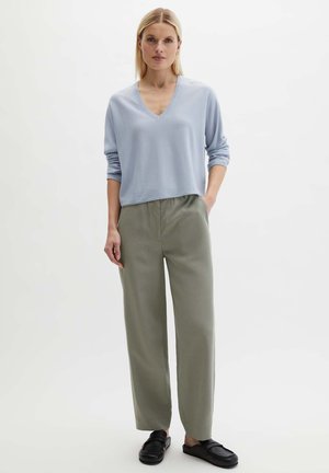 Femme debout, mains dans les poches, portant un pull col V bleu clair, un pantalon large olive, et des chaussures noires sans lacets sur fond blanc.