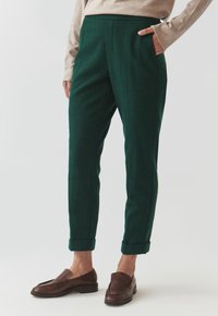 Green, textured wool trousers with a tapered leg, side pockets, and rolled cuffs, paired with dark brown slip-on shoes.