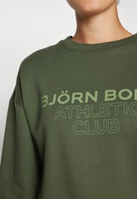 Olajzöld pulóver, kerek nyakkal, laza szabással, elöl világoszöld betűkkel "BJÖRN BORG ATHLETIC CLUB" felirattal.