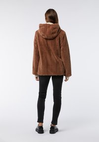 Giacca in finta pelliccia marrone con cappuccio, caratterizzata da un motivo a zigzag testurizzato. Abbinata a pantaloni neri slim-fit e calzature nere.