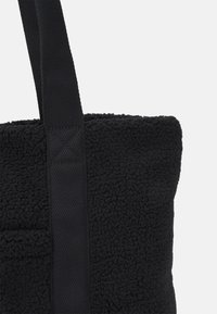 Černá taška z texturované fleecové látky, s pevným tkaným popruhem a jemným detailem přední kapsy. Jednoduchý design.