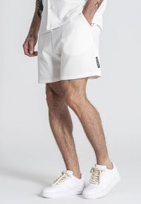 Shorts blancos de textura con un corte holgado, que cuentan con bolsillos y una etiqueta negra en el costado. Se combinan con zapatillas blancas y cordones texturizados.