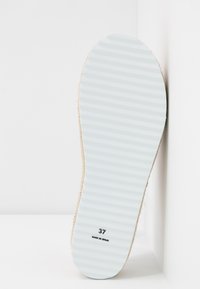 Bílé, žebrované podrážky s texturovaným hnědým okrajem. Velikost 37 a "VYROBENÉ VE ŠPANĚLSKU" vytištěné na spodní straně.