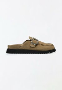 Mocassin marron en suède à enfiler avec semelle noire crantée et détail boucle argentée sur fond blanc uni.