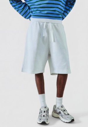 Mężczyzna noszący białe teksturowane krótkie spodenki do kolan ze sznurkiem, białe skarpetki, srebrne sneakersy oraz niebiesko-zieloną, paskowaną, długorękawą koszulkę, stoi.