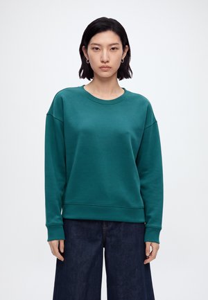 Donna con capelli corti e neri, che indossa orecchini a cerchio dorati, una felpa a maniche lunghe color turchese e jeans scuri a gamba larga, in piedi su sfondo bianco.