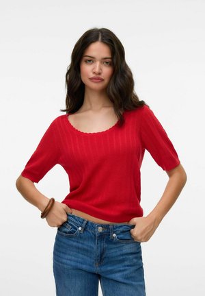 Maglione rosso lavorato a maglia con maniche corte, texture a coste, scollatura rotonda ondulata, indossato con jeans in denim blu.